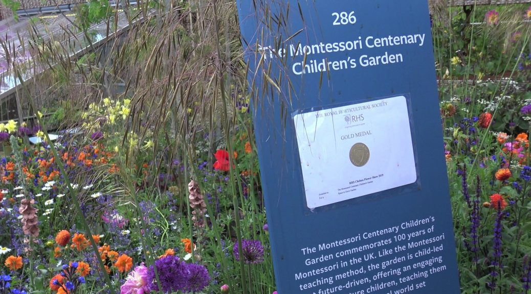 Information plinth with gold medal on in front of The Montessori garden at Chelsea with colourful flowers in the back ground.