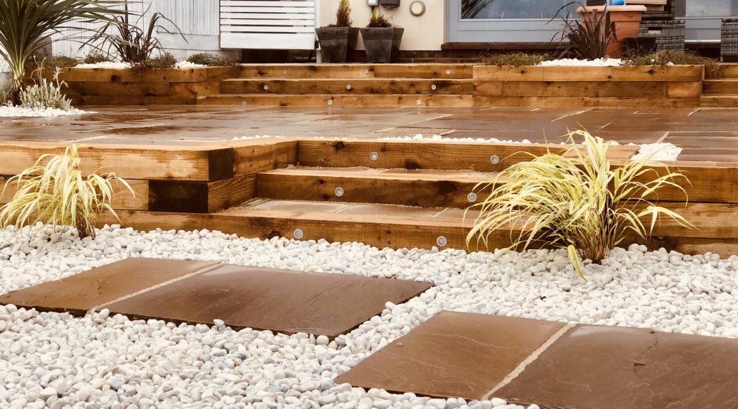 A beach themed garden with steps leading to raised area. all made out of wood. White pebbles in the forefront with patio slabs as stepping stones around them.